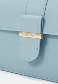 Světle modrá kožená kabelka s klopovým uzávěrem, zlatými kovovými detaily a jemným prošíváním. Hladký povrch a strukturovaný design.