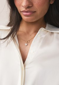 Collier en or avec un pendentif en forme de cœur orné de pierres transparentes, porté sur une chemise blanche boutonnée au tissu lisse.