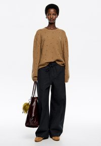 Jersey marrón con adornos negros, combinado con pantalones holgados de color gris oscuro. Accesorios con un bolso burdeos brillante y zapatos beige.