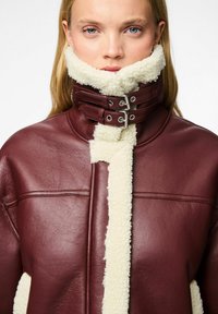 Giacca in pelle bordeaux con collare in shearling crema, collo alto e accessori in argento. Pelle strutturata con cuciture e punti visibili.