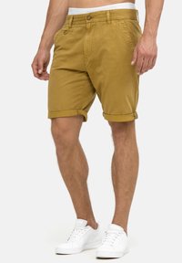 Shorts en coton beige avec des ourlets retroussés, fermeture à bouton à l'avant et poches latérales, portés avec des baskets blanches sur un fond uni.
