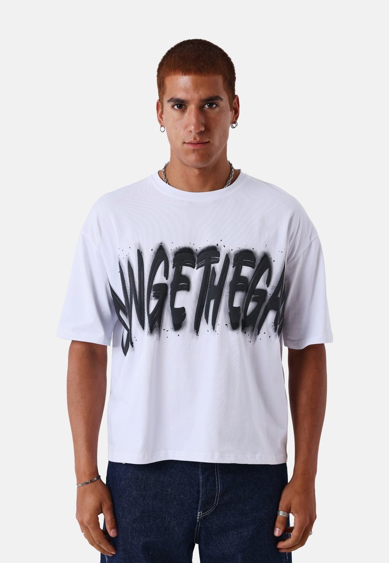 Jeune homme aux cheveux roux courts portant un T-shirt blanc avec un grand texte noir style peinture en spray et un jean bleu, debout devant un fond uni.