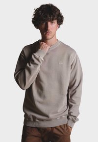 Sudadera de cuello redondo beige hecha de un tejido suave, con puños y bajo acanalados, y un pequeño logo de elefante bordado en el pecho.