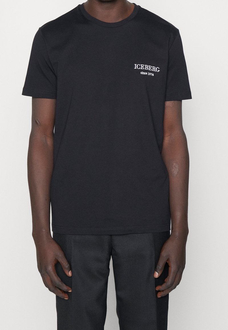 T-shirt noir à manches courtes avec texte blanc "ICEBERG since 1974" porté par une personne à la peau foncée, debout devant un fond clair uni.