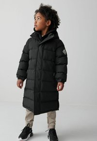 Svart puffig jacka med huva, med quiltade sektioner och sidofickor med dragkedja. Buren över beige byxor och svarta sneakers med vita detaljer.