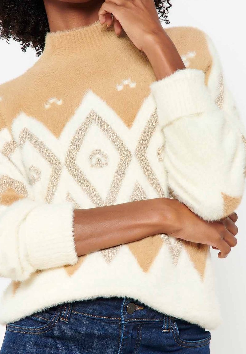 Pull crème et beige en matériau doux, présentant un motif en losanges et des poignets côtelés. Design élégant avec un col montant.