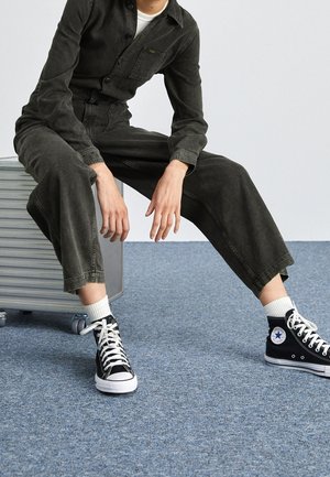 Combinaison en denim noir à manches longues avec une poche frontale, associée à des baskets montantes noires et des chaussettes blanches côtelées. Sol gris.