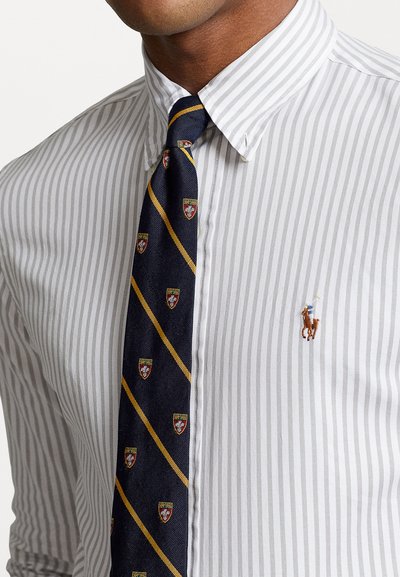 Cămașă cu dungi albe și gri, cu guler cu nasturi. O cravată albastră cu dungi diagonale galbene și modele emblematice.