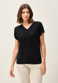 Černý top s krátkým rukávem a výstřihem do V, který má jemnou texturu a středový šev. Stylizovaný s ohrnutými rukávy, spárován se světle zbarvenými kalhotami.