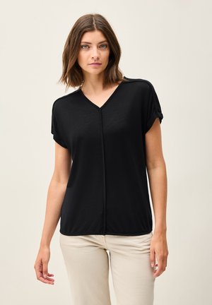 Haut noir à manches courtes avec un décolleté en V, présentant une texture douce et une couture centrale. Stylisé avec des manches retroussées, associé à un pantalon de couleur claire.