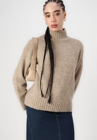 Beige stickad tröja med hög krage och ribbad fåll, parat med en jeanstub. Modellen bär en beige axelväska.