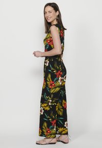 Jumpsuit floreale in tessuto nero, con fiori rossi e bianchi, foglie verdi, vestibilità aderente, design senza maniche e gamba larga. Cinghie alla caviglia argentate visibili.