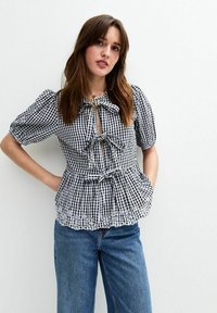 Top a cuadros blanco y negro con mangas abullonadas, frente con ojal y detalles de lazo. Presenta un dobladillo con forma de onda y acentos de ojales. Combinado con jeans azules.
