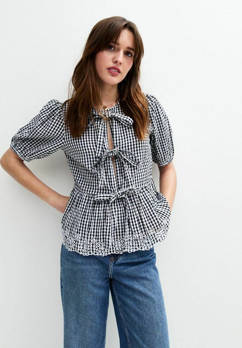 Top a cuadros blanco y negro con mangas abullonadas, frente con ojal y detalles de lazo. Presenta un dobladillo con forma de onda y acentos de ojales. Combinado con jeans azules.