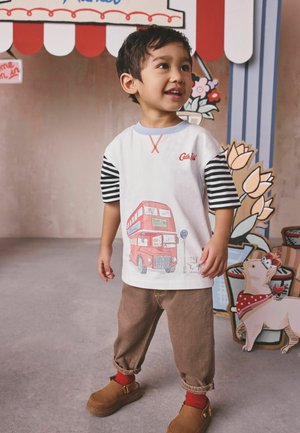 Junger Junge, der lächelt und steht, trägt ein weißes Hemd mit schwarzen gestreiften Ärmeln und einer Grafik eines roten Doppeldeckerbusses, braune Hosen und braune Schuhe.