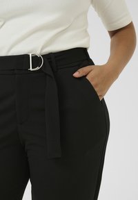 Femme portant un pantalon noir avec une ceinture en tissu et une boucle en D argentée, main dans la poche, et un haut blanc à manches courtes.