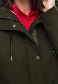 Chaqueta acolchada verde oliva con cierre de cremallera y botones, que presenta una sudadera roja debajo y costuras de acento negras.