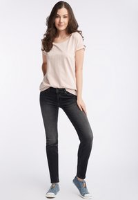 Jasnoróżowa koszulka z krótkim rękawem z haftowanym napisem, zestawiona z dopasowanymi czarnymi dżinsami i niebieskimi trampkami.