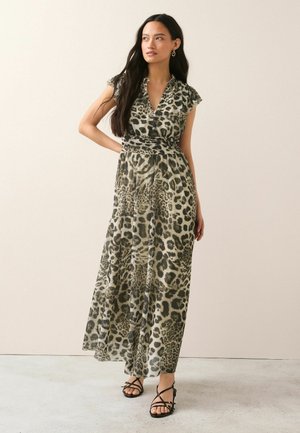 Kobieta o długich ciemnych włosach, ubrana w bezrękawnikową maxi sukienkę w panterkę i czarne sandały na paski, stoi na tle jednolitego tła.