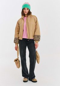 Žena v béžovej puffovej bunde s leopardími manžetami, ružovom svetri, čiernych džínsach, neónovo zelenom a ružovom čiapke, drží zodpovedajúce béžové tašky.