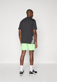 T-shirt de sport noir avec des rayures blanches dans le dos, associé à un short vert vif. Le modèle porte des baskets noires et des chaussettes blanches.