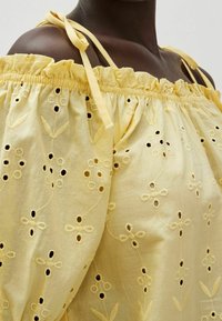 Lys gul, off-the-shoulder bluse med flæset halsudskæring, broderede blomster mønstre og hulmønster detaljer; har bindebånd og blød tekstur.