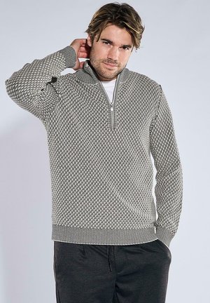 Mann trägt schwarz-weiß gemusterten Reißverschluss-Pullover mit Hand hinter dem Nacken und dunkle Hose vor einfarbigem Hintergrund.