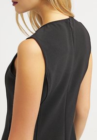 Haut noir sans manches avec une texture lisse, présentant un col montant et une fermeture éclair dans le dos, conçu avec des lignes épurées et des accents minimaux.