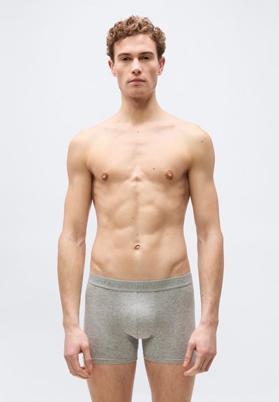 Νεαρός άνδρας με σγουρά μαλλιά που φοράει ανοιχτό γκρι boxer briefs Polo Ralph Lauren, στέκεται μπροστά σε απλό ανοιχτό φόντο.