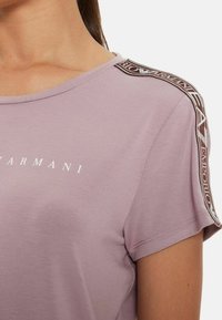 Levandulové bavlněné tričko s kulatým výstřihem. Má logo na přední straně a vzorovaný akcent podél ramenního švu. Krátké rukávy.