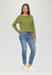 Groene lange mouw trui met een bootnek, gecombineerd met blauwe distressed skinny jeans en witte sneakers, staande tegen een neutrale achtergrond.