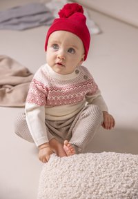 Strickpullover mit rotem Fair-Isle-Muster und cremefarbenen Ärmeln, graue Ripp-Hosen und eine rote Strickmütze mit Pompon, die auf einer hellen Oberfläche sitzt.