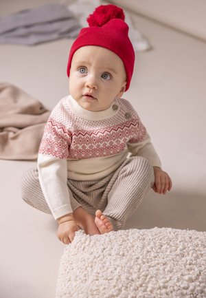 Maglione lavorato a maglia con motivo fair isle rosso e maniche beige, pantaloni grigi a coste e un berretto lavorato a maglia rosso con pompon, posizionato su una superficie chiara.