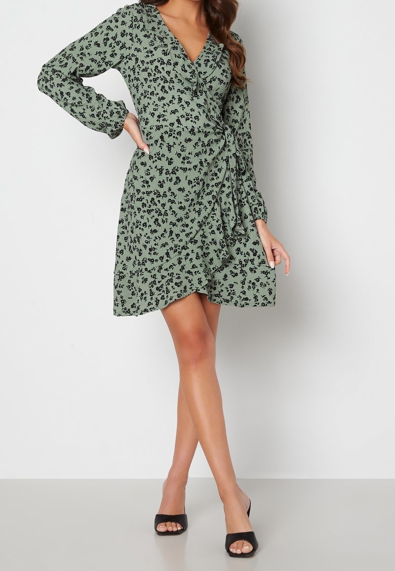 Femme portant une robe portefeuille verte à imprimé floral noir et manches longues, debout avec une main sur la hanche et des talons ouverts noirs.