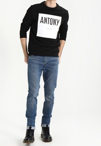 Sudadera negra con un gráfico rectangular blanco que dice "ANTONY ITALIA". Llevada con jeans azules y botas negras con cordones, que tienen los dobladillos acampanados.