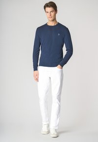 Maglione blu navy a maniche lunghe realizzato in tessuto a maglia, con orlo e polsini a coste. Indossato con pantaloni bianchi e sneaker di colore chiaro.