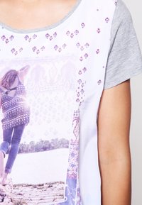 Grafické tričko má bílé tělo a šedé rukávy, na kterých je barevný fotografický potisk a složité vzory v purpurových a růžových tónech.