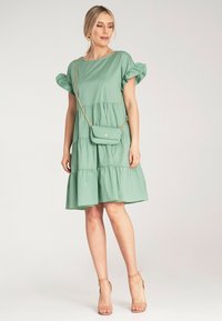 Jasnozielona sukienka warstwowa z bufkami, wykonana z miękkiego materiału. Uzupełniona małą torebką i neutralnymi sandałami na obcasie.