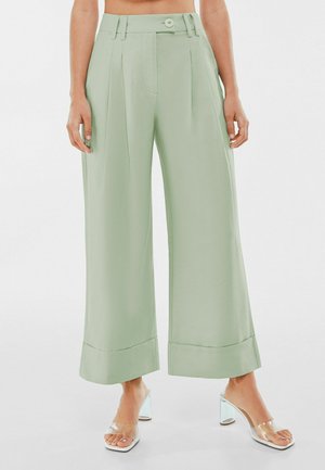 Pantalon large taille haute vert menthe avec plis, ourlets retroussés et fermeture à bouton, porté avec des sandales à talons transparentes.