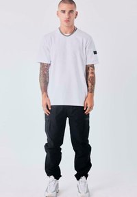 Joven con pelo corto y tatuajes lleva una camiseta blanca, pantalones cargo negros, zapatillas blancas y cadenas de plata, de pie contra un fondo blanco.