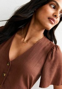 Blusa marrón de botón con mangas cortas abullonadas, que presenta un escote en V profundo y una textura ligera. Collares de oro con un pequeño colgante.