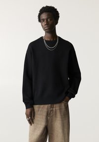 Pulover negru din tricot cu mâneci lungi, având guler rotund și manșete cu aspect texturat, asortat cu pantaloni bej, ușor texturizați. Colier din lant argintiu.