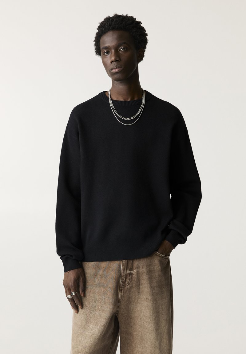Pulover negru din tricot cu mâneci lungi, având guler rotund și manșete cu aspect texturat, asortat cu pantaloni bej, ușor texturizați. Colier din lant argintiu.