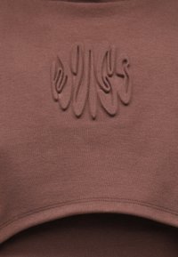 Rjava kratka jopica z reliefnim vzorcem na prsih, mehka tkanina in gladek, okrogel izrez. Brez vidnih zapenjanj.