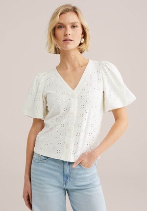 Blonde Frau trägt eine weiße Bluse mit Lochmuster, Knöpfen und kurzen, puffigen Ärmeln sowie hellblaue Jeans, posiert mit einer Hand in der Tasche.