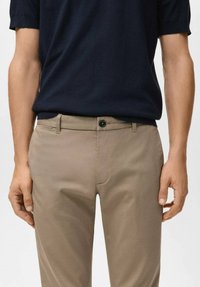 Pantaloni color sabbia in mélange di cotone con una texture liscia, dotati di chiusura con un solo bottone e passanti per cintura. Indossati con una camicia a maniche corte di un blu navy scuro.