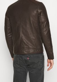 Giubbotto da moto in pelle marrone con un design aderente, pannelli texturizzati sulle spalle e una finitura liscia. Abbinato a jeans in denim scuro.