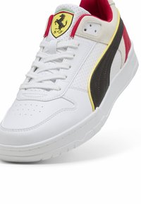 Hvide sneakers med perforeret læderoverdel, røde og sorte detaljer, gul logo på tungen, flad gummisål og snørebåndsdesign.