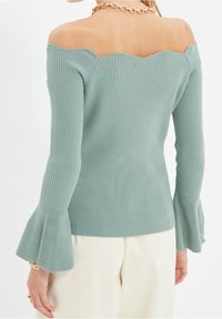 Gerippter, schulterfreier Pullover in gedämpftem Grün mit einem wellenförmigen Ausschnitt und ausstellenden Ärmeln, gefertigt aus weichem Strickmaterial.