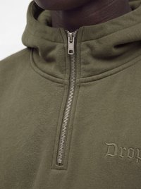 Gros plan d'une personne portant un sweat à capuche zippé vert olive avec une fermeture éclair en métal et un texte partiellement brodé "Drop" sur la poitrine.